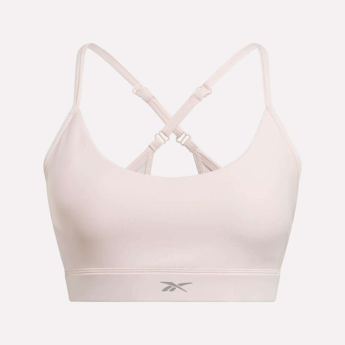The Reebok Women's Lux Strappy Sports Bra in light pink features thin, adjustable crisscross back straps, medium support, a seamless and sweat-wicking design, and a small logo on the front underband. Displayed on a plain white background.