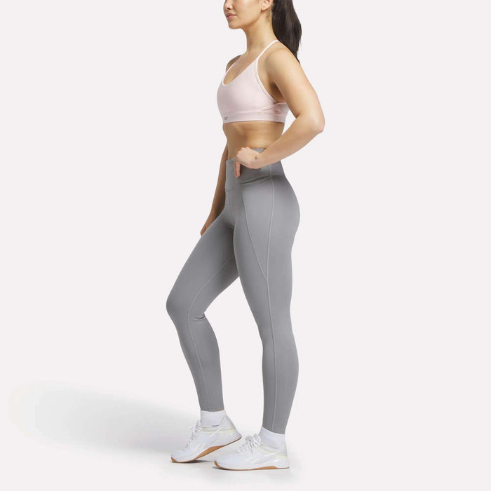 A woman stands in profile against a white background wearing the Reebok Women's Lux Strappy Sports Bra in light pink, paired with high-waisted gray leggings and white sneakers. One hand rests on her hip as she looks ahead.