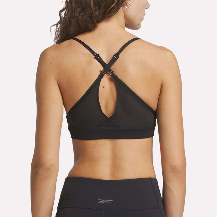 A woman with long, wavy brown hair stands with her back to the camera, wearing Reebok's Women's Lux Strappy Sports Bra featuring adjustable crossed straps and a keyhole back detail, paired with black athletic leggings.