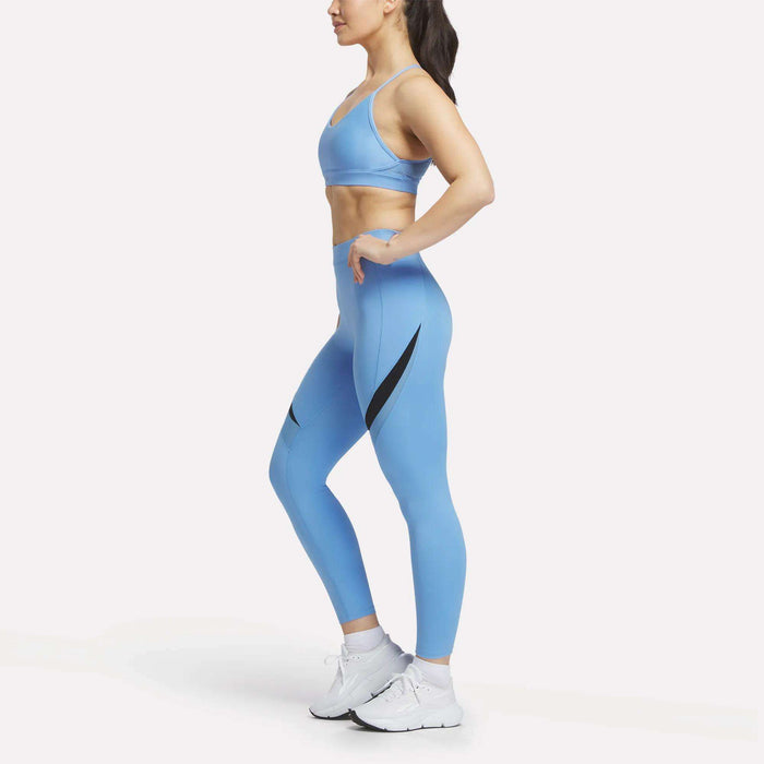 A woman stands in profile with hands on hips, wearing the Reebok Women's Lux Strappy Sports Bra and matching leggings with black accents, plus white sneakers, all set against a plain white background.