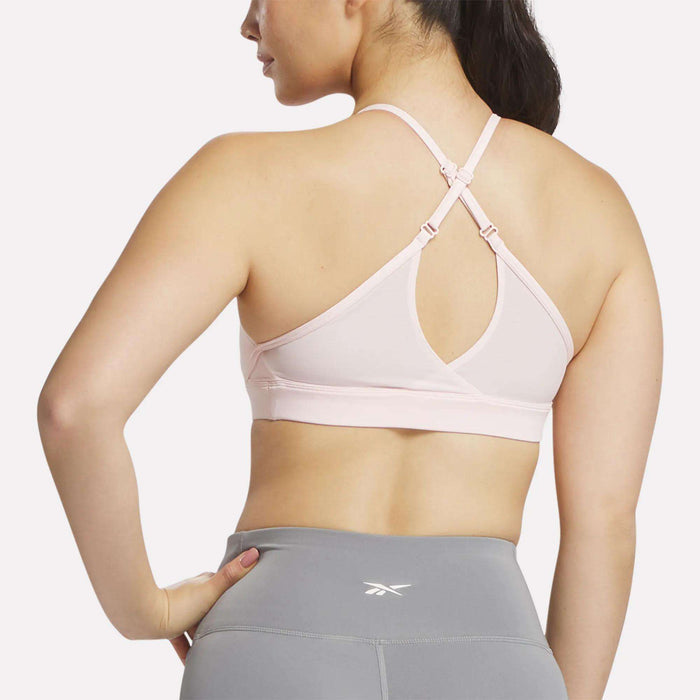 A woman wears the Reebok Women's Lux Strappy Sports Bra in light pink, featuring sweat-wicking fabric and crisscross straps, paired with gray leggings, shown from the back against a plain white background.