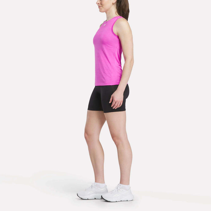 A woman stands in profile against a white background, wearing the Reebok Women's Marled Performance Tank Top in pink with black shorts and white sneakers—highlighting fashionable activewear ideal for workouts.