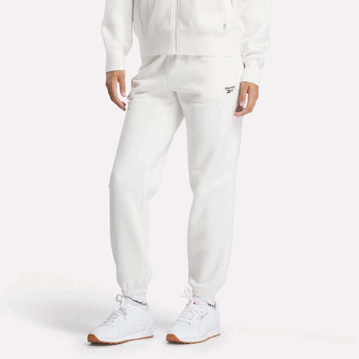 A person wearing Reebok Women's Medium Vector Sweatpants with the logo on the left thigh stands against a plain light background, paired with a white zip-up hoodie and white sneakers.