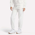 A person wearing Reebok Women's Medium Vector Sweatpants with the logo on the left thigh stands against a plain light background, paired with a white zip-up hoodie and white sneakers.