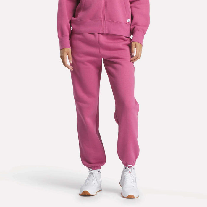 A person stands against a white background wearing Reebok Women's Medium Vector Sweatpants in pink, paired with a matching zip-up hoodie and white Reebok athletic shoes.