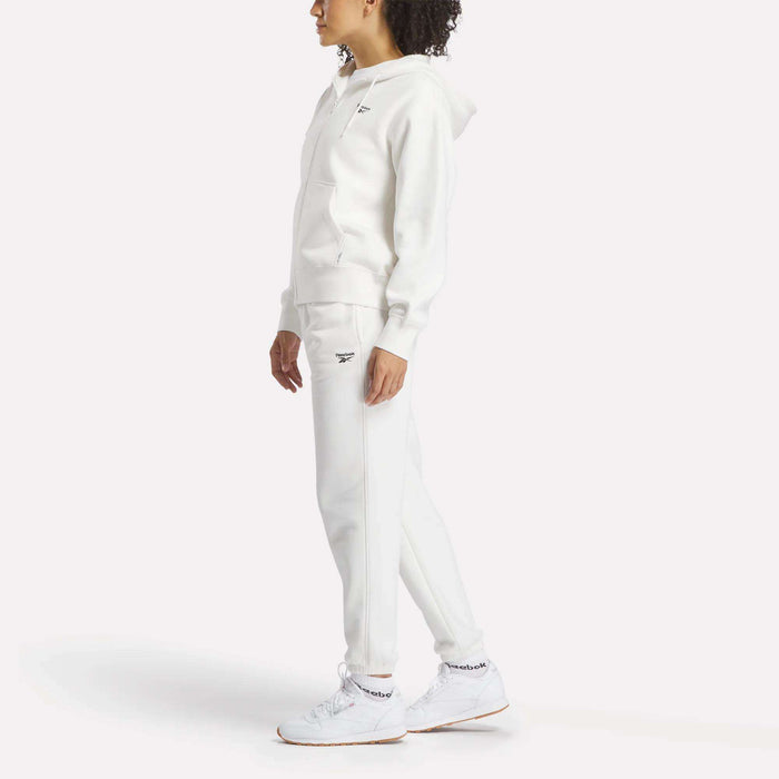 A person stands against a white background wearing an all-white Reebok hoodie, Women's Medium Vector Sweatpants with a small logo, and sneakers. The cotton-blend sweatpants are by Reebok, and the person's face is turned slightly away.