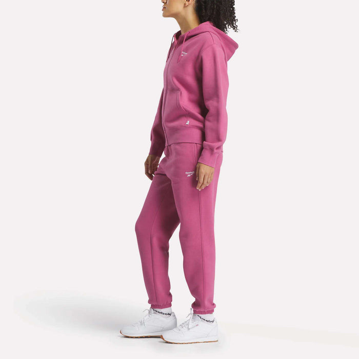 A person wears Reebok Women's Medium Vector Sweatpants with a matching pink hoodie and white sneakers, standing against a plain white background and facing slightly to the left.