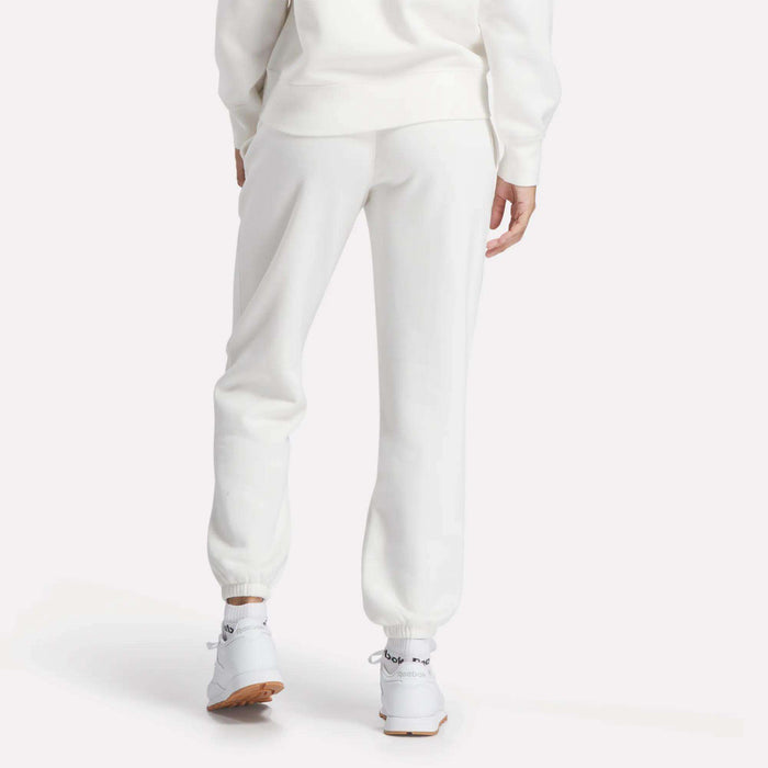 A person stands with their back to the camera wearing Reebok Women's Medium Vector Sweatpants and a matching white sweatshirt. The scene features Reebok sneakers and a plain, light-colored background.