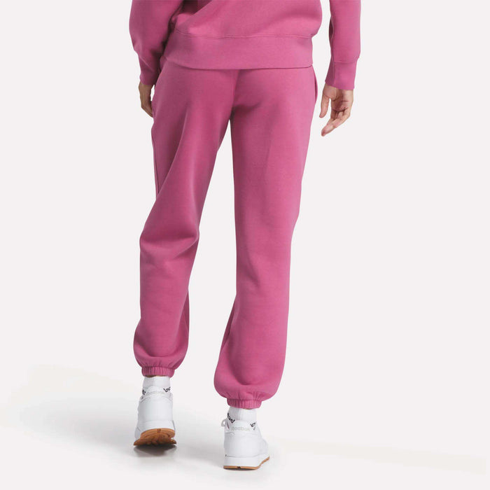 A person faces away in Reebok Women's Medium Vector Sweatpants and a matching sweatshirt, bright pink in color, paired with white sneakers against a plain white background.