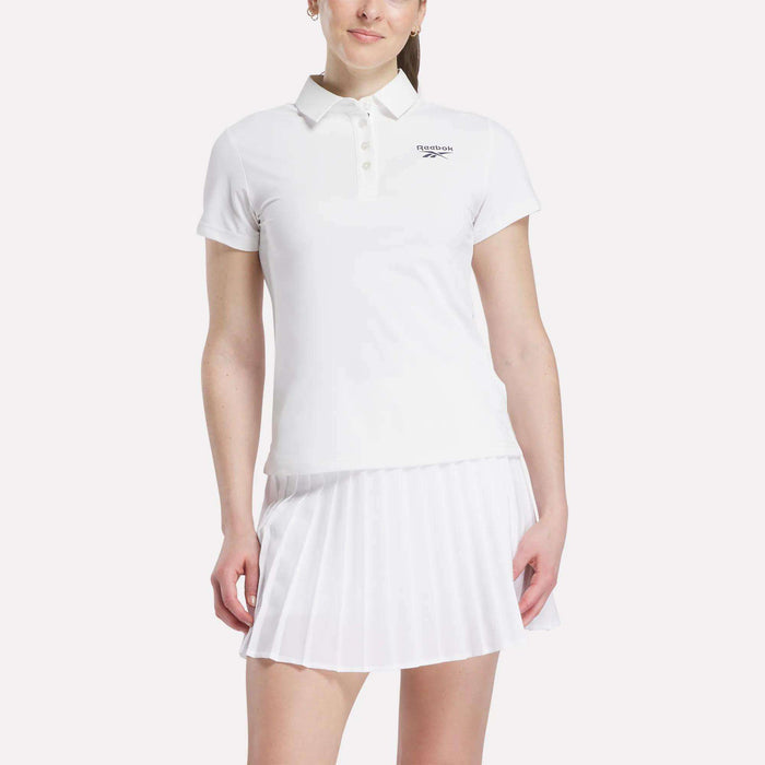 A woman in a white Reebok Women's Micro Pique Polo Shirt with a collar and short sleeves, paired with a white pleated skirt, stands against a plain light background.