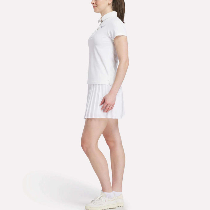 A woman stands in profile wearing a Reebok Women's Micro Pique Polo Shirt, a white pleated skirt, and white sneakers against a plain light background.