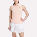 A woman poses confidently in a Reebok Women's Micro Pique Sleeveless Polo Shirt and a white pleated skirt, highlighting the sporty look of this UPF-rated outfit against a plain backdrop.