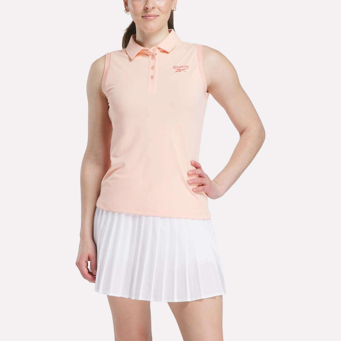 A woman poses confidently in a Reebok Women's Micro Pique Sleeveless Polo Shirt and a white pleated skirt, highlighting the sporty look of this UPF-rated outfit against a plain backdrop.