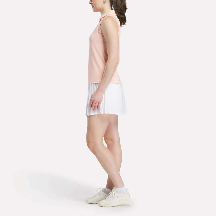 A woman in profile wears a Reebok Women's Micro Pique Sleeveless Polo Shirt in peach, paired with a white pleated skirt and white sneakers, set against a plain white background.