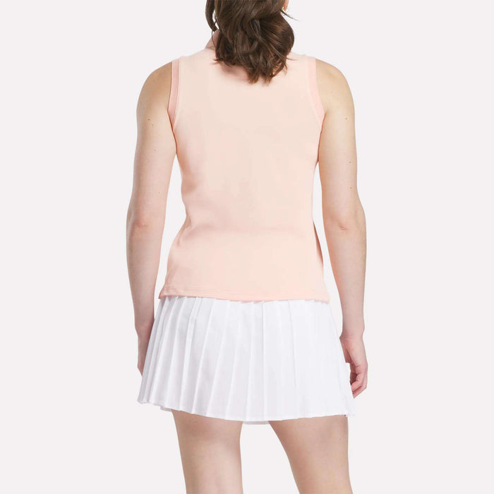 A woman with partially tied-back brown hair faces away, wearing a peach Reebok Women's Micro Pique Sleeveless Polo Shirt and a white pleated skirt. The plain white background highlights the polo's UPF protection.