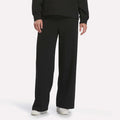 A person wearing Reebok Women’s Mid-Rise Wide-Leg Pants in black, white shoes, and a long-sleeve black top stands against a plain white background.