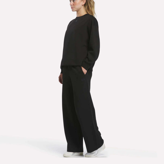 A person stands in profile against a plain white background, wearing a loose black sweatshirt and Reebok Women’s Mid-Rise Wide-Leg Pants with hands in pockets, paired with white sneakers.