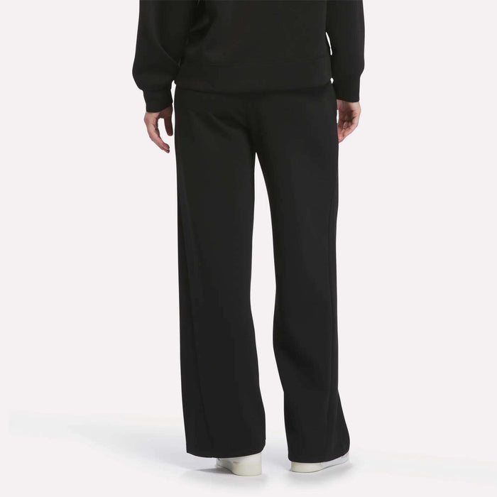 A person is seen from behind against a plain light background, wearing Reebok Women’s Mid-Rise Wide-Leg Pants in black with a relaxed fit, paired with a black top and white shoes.