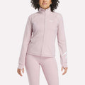 A person wearing a light pink Reebok Women's Motion Core Full-Zip Hoodie and matching leggings, standing with arms relaxed by their sides against a plain white background.