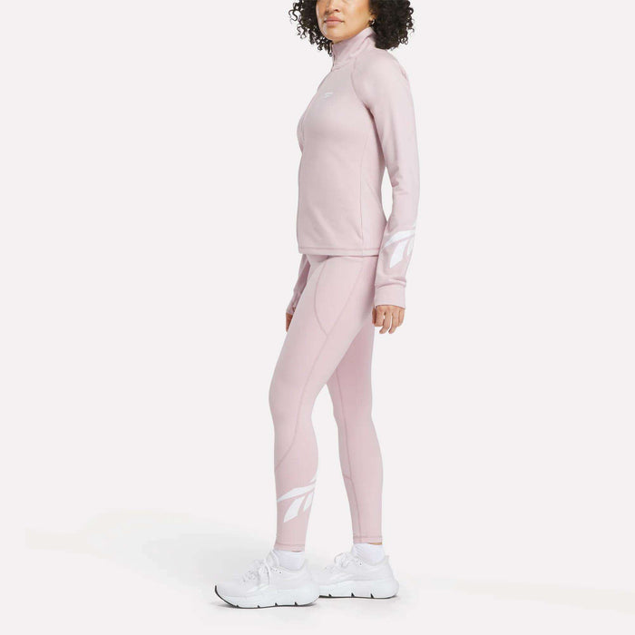 A woman stands in side profile against a white background wearing the Reebok Women's Motion Core Full-Zip Hoodie in light pink, styled with matching athletic pants and white sneakers. The hoodie features bold white logo designs on the sleeves and lower legs.