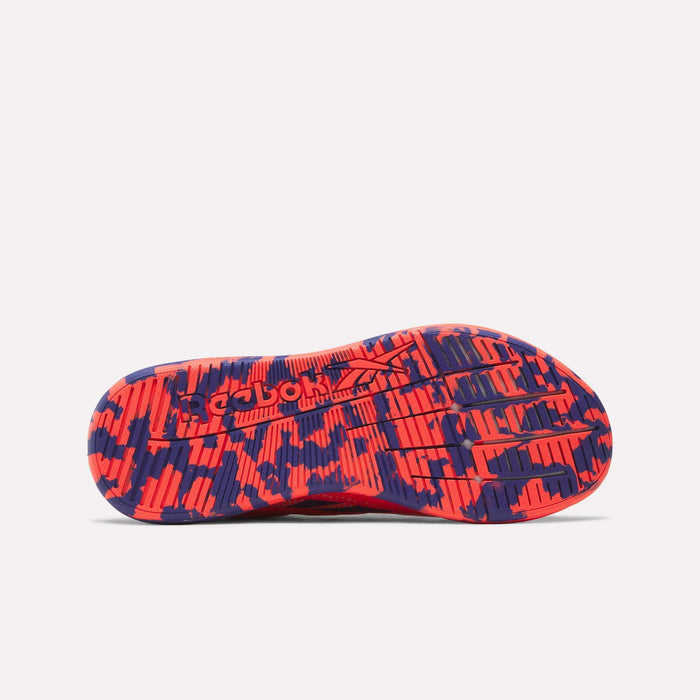 The image features the sole of a women's Reebok Nano X5 Edge Training Shoe with a striking red and navy camo pattern. The Reebok logo appears in the center, while grooved lines and textured areas deliver grip for intense training sessions.