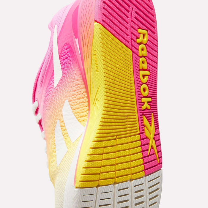 Close-up of Reebok Women's Nano X5 Training Shoes, featuring a pink, yellow, and white gradient design. The comfortable sole displays bold yellow Reebok branding and textured grip for improved traction.
