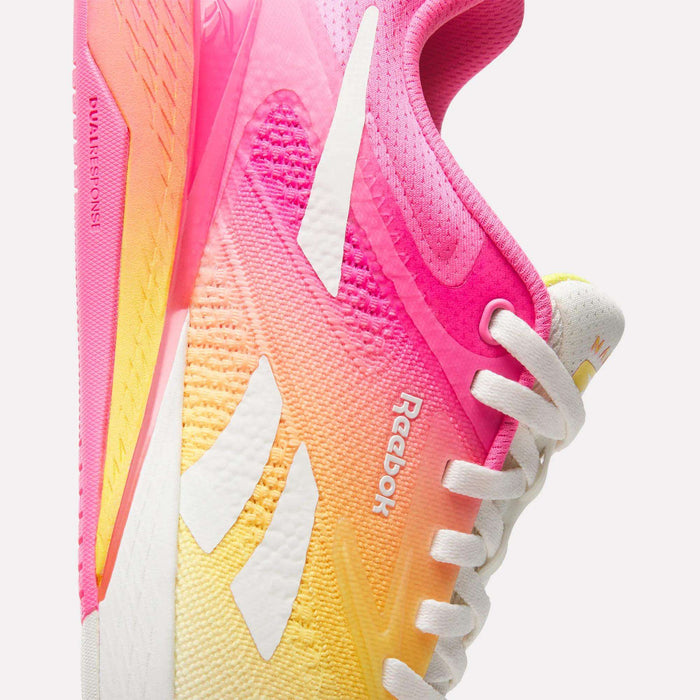 Close-up of Reebok Women's Nano X5 Training Shoes in a pink, yellow, and white gradient. This athletic shoe has white laces, the Reebok logo, a textured sole, and mesh for breathability and support.