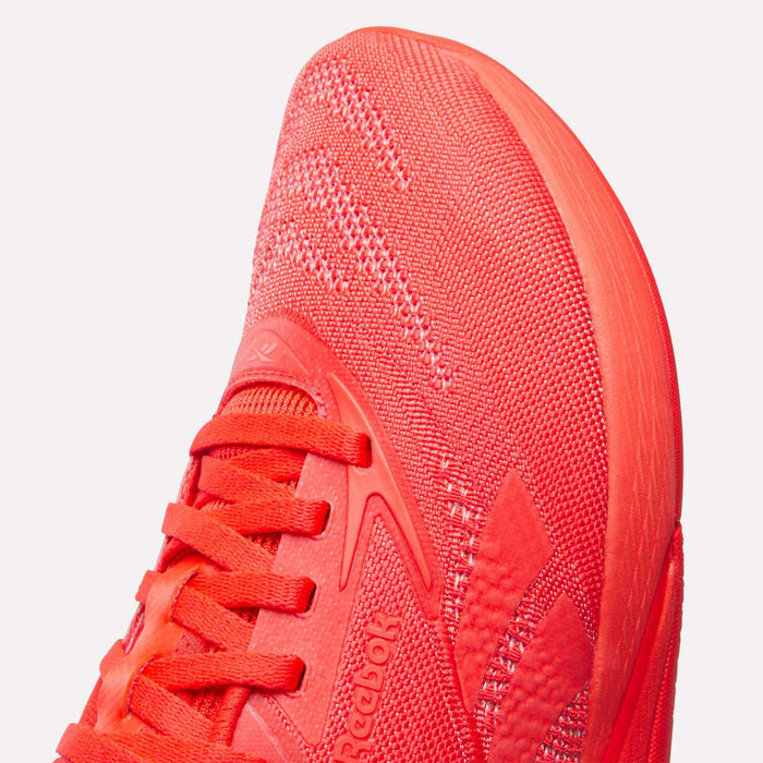 Close-up of the front and side of Reebok Women's Nano X5 Training Shoes in bright red, showcasing a textured knit upper, laces, visible Reebok branding on the side, with detailed sole and stitching.