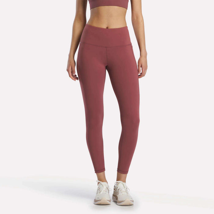 A woman stands facing forward in Reebok Women's Odyssey High-Rise 7/8 Leggings in Blackberry, paired with a matching sports bra and white athletic shoes, arms relaxed at her sides against a plain white background.