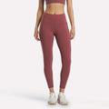 A woman stands facing forward in Reebok Women's Odyssey High-Rise 7/8 Leggings in Blackberry, paired with a matching sports bra and white athletic shoes, arms relaxed at her sides against a plain white background.