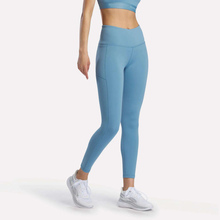A woman wears Reebok Women's Odyssey High-Rise 7/8 Leggings in light blue with a matching sports bra and white athletic shoes, standing with one arm relaxed against a plain white background.