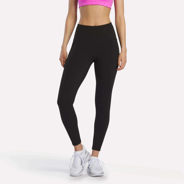A person models Reebok Women's Odyssey High-Rise 7/8 Leggings and a bright pink sports bra with white athletic shoes, standing against a plain white background. The model’s head is not visible in the image.