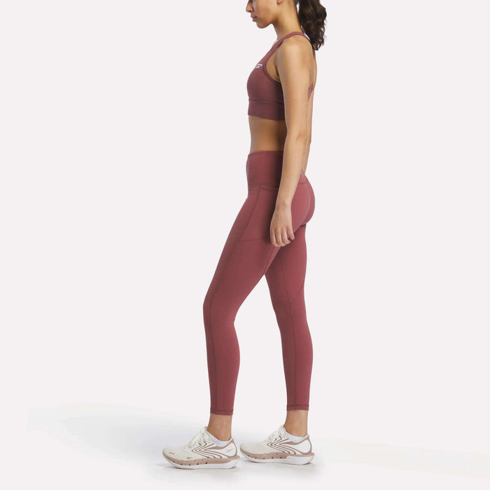 A woman in profile faces left, wearing Reebok Women's Odyssey High-Rise 7/8 Leggings and a matching sports bra, paired with white athletic shoes, set against a plain white background.