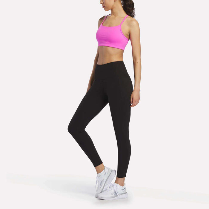 A woman stands sideways in a bright pink sports bra, Reebok Women's Odyssey High-Rise 7/8 Leggings, and white athletic shoes against a plain white background.