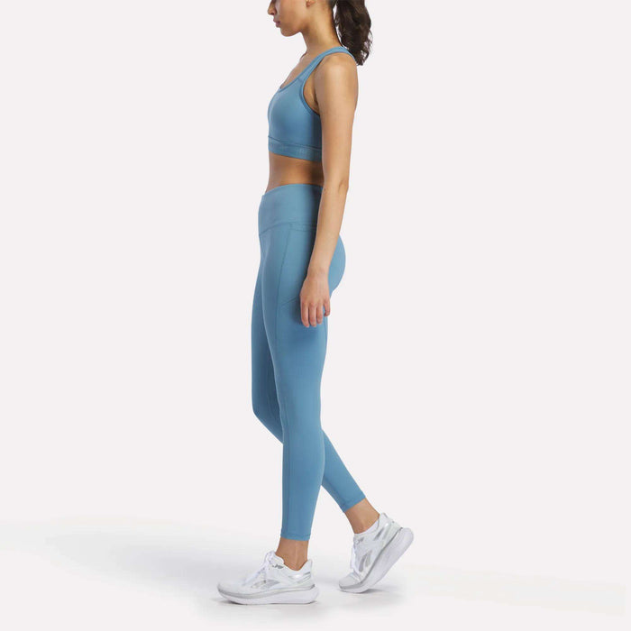 A woman in Reebok Women's Odyssey High-Rise 7/8 Leggings, paired with a sports bra and white running shoes, stands in profile against a plain white background.