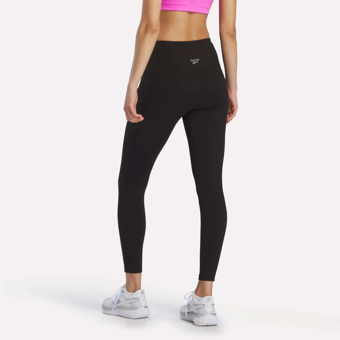 A woman faces away from the camera, modeling bright pink Reebok Women's Odyssey High-Rise 7/8 Leggings and white athletic sneakers, standing against a plain white background.