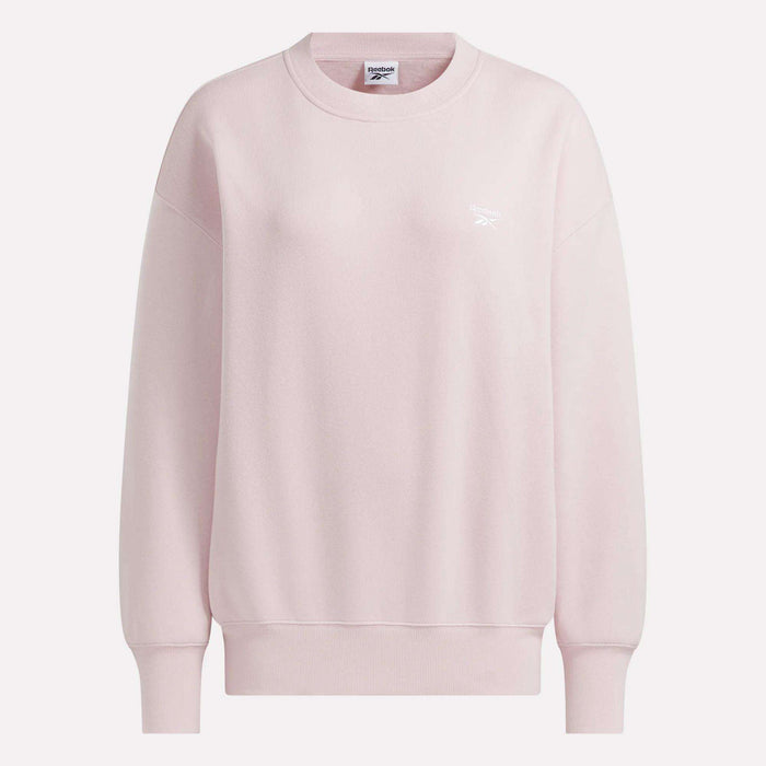 The Women’s Oversized Crewneck Sweatshirt by Reebok is shown in light pink, featuring long sleeves, ribbed cuffs and hem, and a small white Reebok logo on the left chest. This comfortable sweatshirt is displayed against a plain light background.