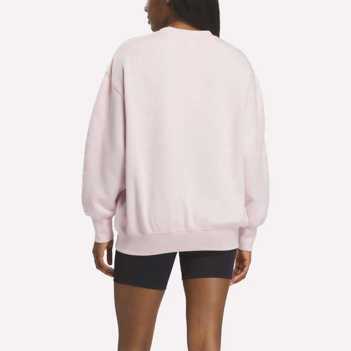 A person stands facing away from the camera against a plain white background, wearing a light pink Reebok Women’s Oversized Crewneck Sweatshirt and black shorts.