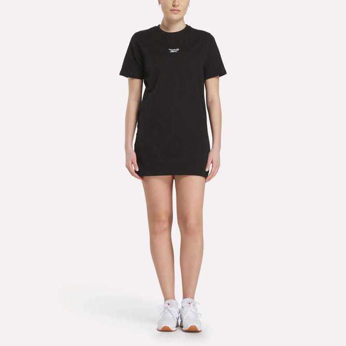 A person faces forward in a Women's Oversized T-Shirt Dress by Reebok—black with a small white chest logo—paired with white sneakers, against a plain white background.