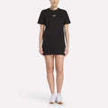 A person faces forward in a Women's Oversized T-Shirt Dress by Reebok—black with a small white chest logo—paired with white sneakers, against a plain white background.