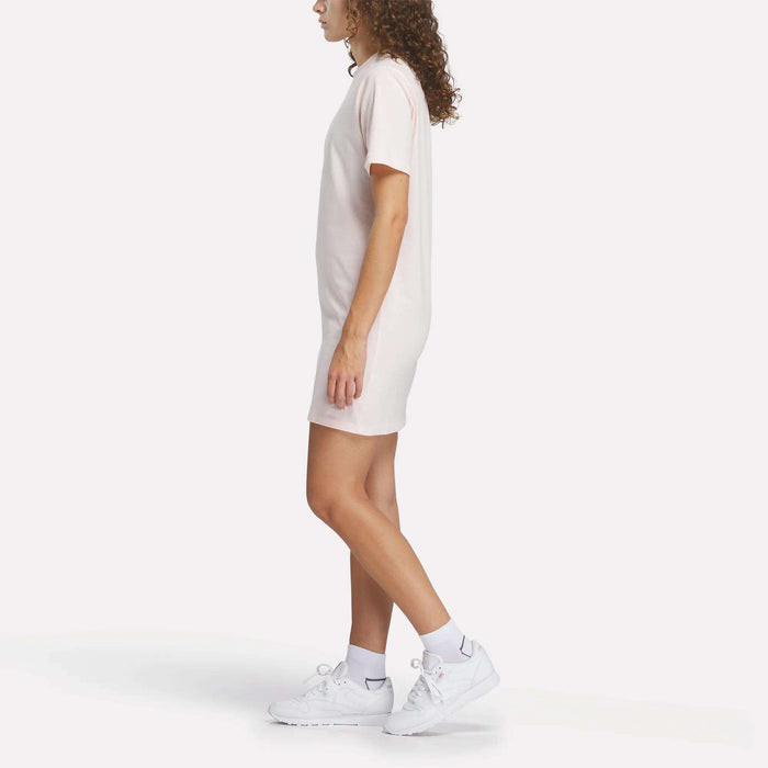 A woman with curly hair stands in profile against a white background, wearing the Reebok Women’s Oversized T-Shirt Dress in light pink, paired with white ankle socks and Reebok sneakers.