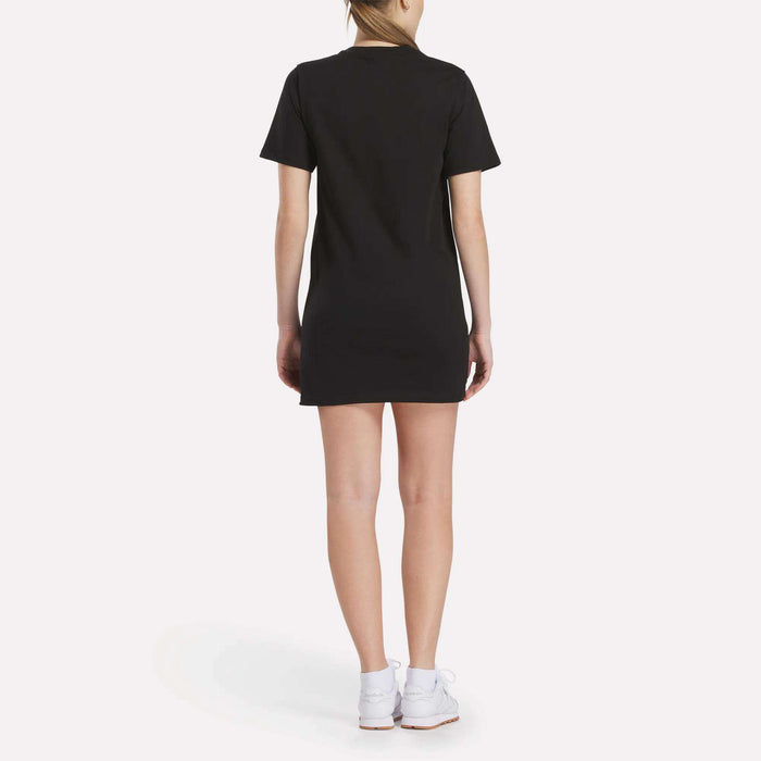 A person faces away, wearing the Reebok Women's Oversized T-Shirt Dress in black with white sneakers, set against a plain white background.