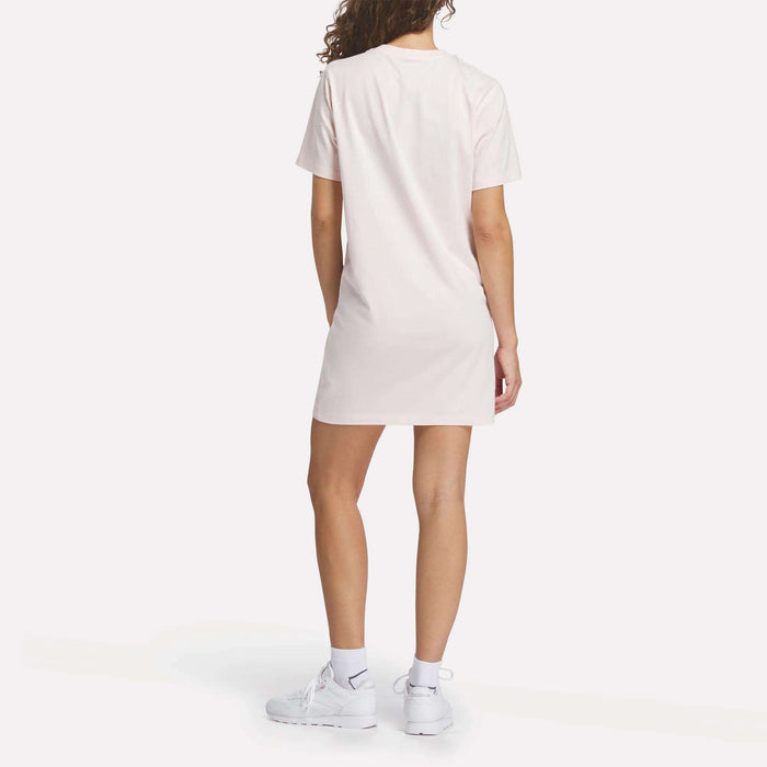 A person faces away, wearing the Reebok Women's Oversized T-Shirt Dress in light pink with white socks and Reebok sneakers, set against a plain white background.
