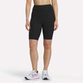 A person is shown from the shoulders down against a white background, wearing Reebok Women's Peached Biker Shorts 9" in black, paired with white athletic sneakers featuring pink accents.