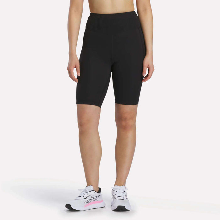 A person is shown from the shoulders down against a white background, wearing Reebok Women's Peached Biker Shorts 9" in black, paired with white athletic sneakers featuring pink accents.