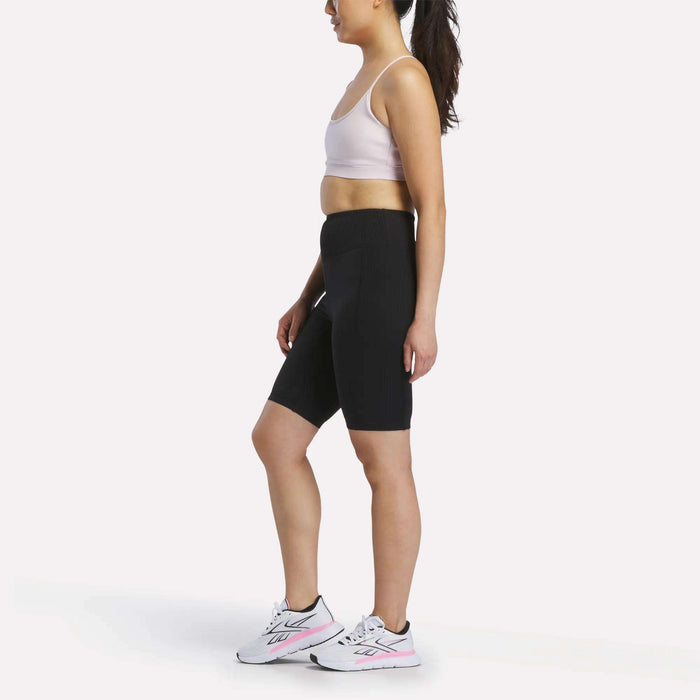 A woman stands in profile against a plain white background, wearing a light sports bra, Reebok Women's Peached Biker Shorts 9", and white athletic shoes with pink accents.