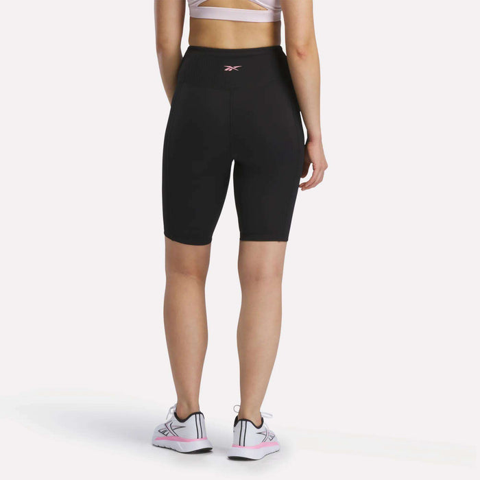 A person stands facing away against a plain white background, wearing a white sports bra, Reebok Women's Peached Biker Shorts 9" in black made from peached fabric, and white sneakers with pink accents, showing the back view of the outfit.