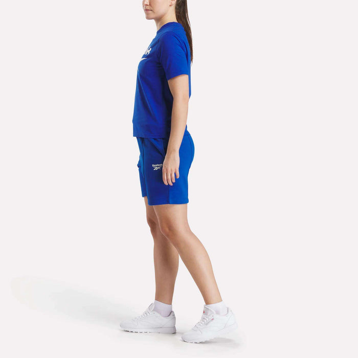A woman in a ponytail stands sideways against a white background, wearing Reebok Women's Pintuck Long Shorts paired with a matching blue cotton-blend fleece t-shirt and white athletic shoes.