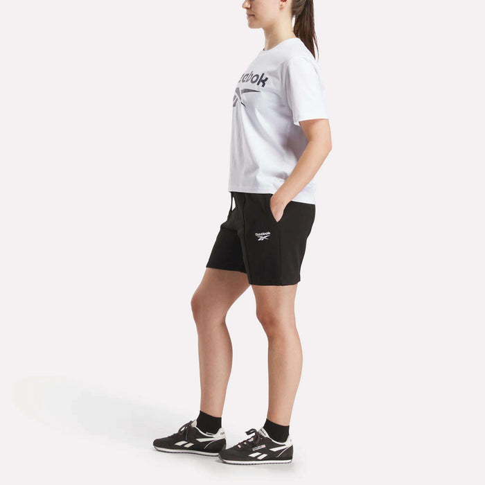 A woman stands sideways against a plain white background wearing a white Reebok t-shirt, black Women’s Pintuck Long Shorts by Reebok with the logo, black socks, and black sneakers.