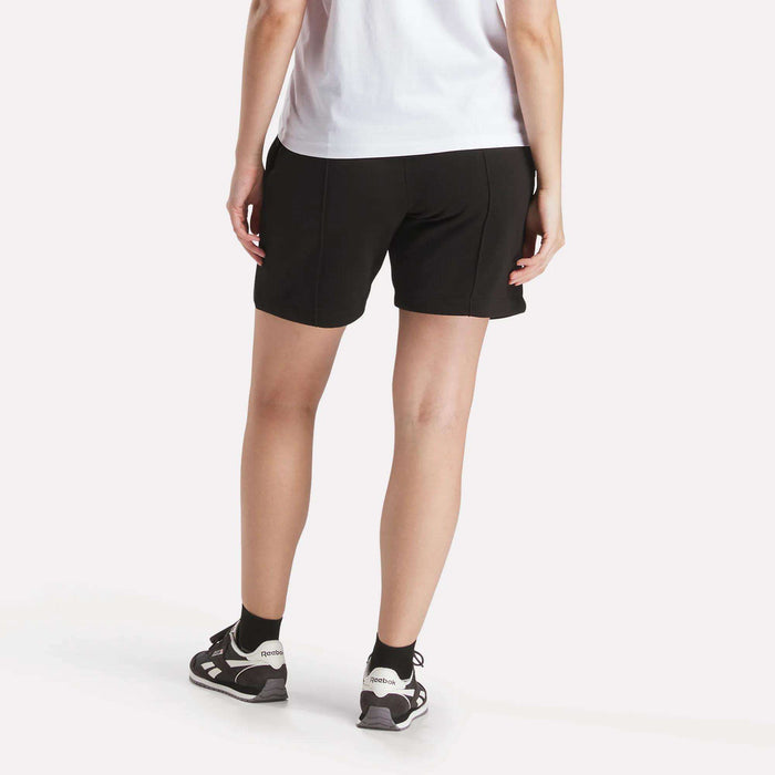A person faces away from the camera in a white t-shirt and Reebok Women's Pintuck Long Shorts, styled with black socks and black-and-white athletic shoes, set against a plain white background.
